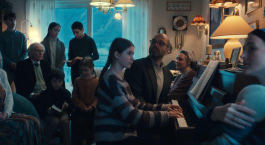 Still from Fjord: a group of people gather around a piano in a warmly lit, wood-panelled room.