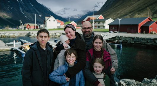 Still from Fjord: a family of six pose together in front of a fjord village with red and white buildings and mountains.