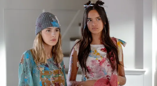 Still from Freakier Friday: two women, one in a patterned jacket and a woollen hat and one with long dark hair in a floral top, stand in a bright hallway.