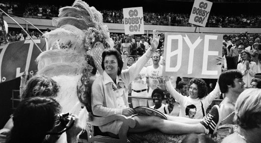 Black-and-white photograph of a crowd of spectators at a tennis event, some holding placards, with a banner behind them.