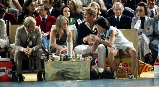 Photograph from Give Me the Ball: a woman in a white tennis outfit sits courtside among a row of seated spectators, with drinks on a table in front of her.