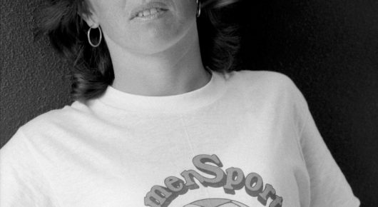 Close-up black-and-white photograph of a woman in a white T-shirt with partly visible printed lettering and a hoop earring.