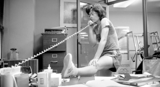 Black-and-white photograph of a woman sitting on a desk in an office with her foot raised, talking on a telephone with a long curly cord.