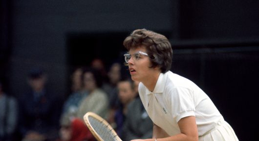 Photograph from Give Me the Ball: a tennis player with glasses and dark hair, in a white dress, crouches ready with a racket, with spectators behind her.