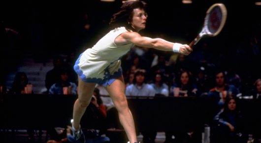Photograph from Give Me the Ball: a tennis player in a white top and blue shorts stretches low to hit a shot, with spectators in the dark behind her.