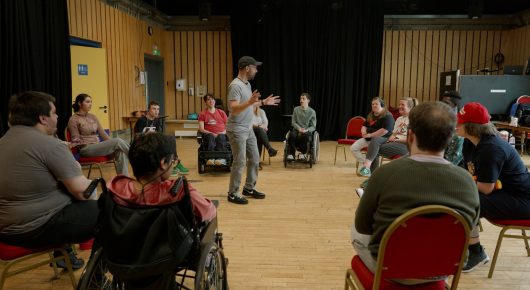 Still from Cambridge Connections: a group of people sit in a circle in a rehearsal hall, with a person standing at the centre.