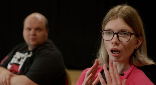 Still from Cambridge Connections: a man in a black T-shirt sits beside a woman with glasses and long hair who clasps her hands, in front of a black curtain.
