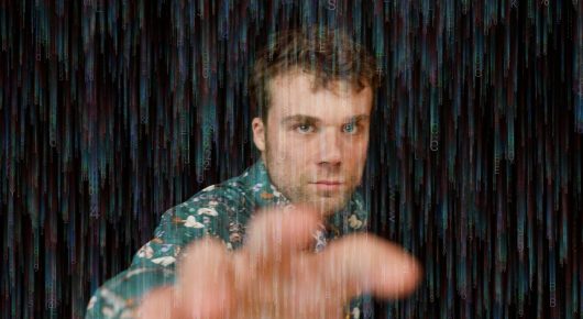 Still from Cambridge Connections: a man in a patterned shirt reaches a hand towards the camera against a dark background streaked with blue and green light.