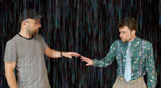 Still from Cambridge Connections: two men in patterned and striped tops reach out and touch hands in front of a dark background streaked with rain-like light.