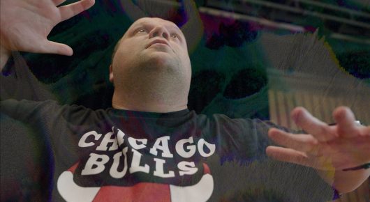 Still from Cambridge Connections: a man in a Chicago Bulls T-shirt lies on his back with his hands raised, in distorted purple light.