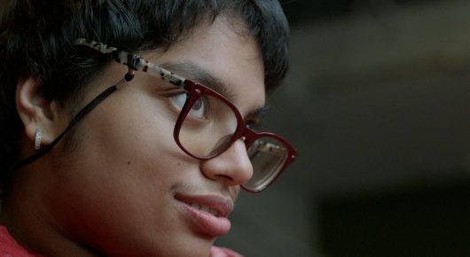 Still from Cambridge Connections: a close-up of a young person with glasses and short dark hair looking to one side.