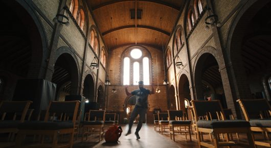 Still from Grappling Grace: a dancer stands in the aisle of a large church with wooden pews and a tall arched ceiling.