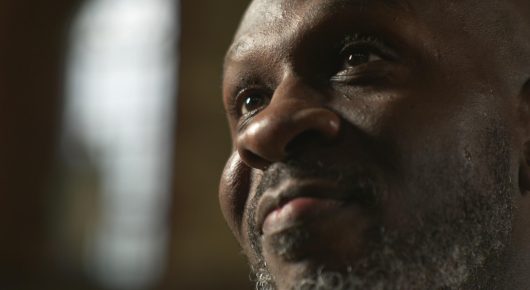 Still from Grappling Grace: a close-up of a man with a short grey beard looking up and to one side.