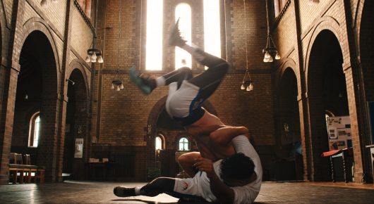 Still from Grappling Grace: a dancer is upside down mid-move above another person on the floor of a church with high arches.