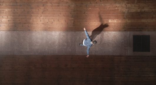 Still from Grappling Grace: a dancer in white is caught mid-air in front of a dark brick wall.