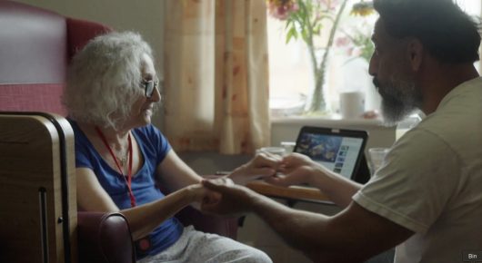 Still from Grappling Grace: an older woman with white curly hair and a bearded man sit close together holding hands, with a laptop between them and a suitcase beside them.