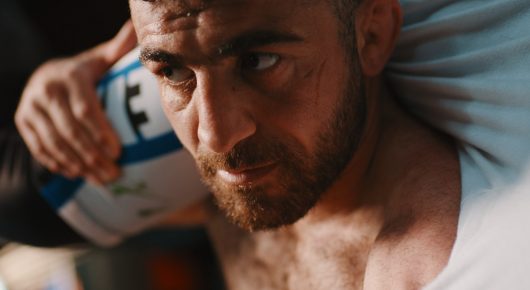 Still from Grappling Grace: a close-up of a bearded man lying back on a pillow with a ball in his hands.
