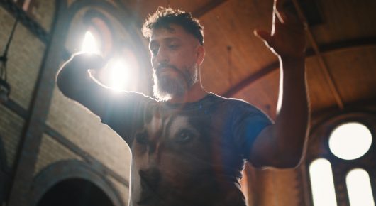Still from Grappling Grace: a man in a blue patterned T-shirt raises one hand in a church, with bright light behind him.