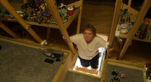 Still from Grappling Grace: a boy looks up from an open hatch in the floor of a loft filled with toys and shelves.