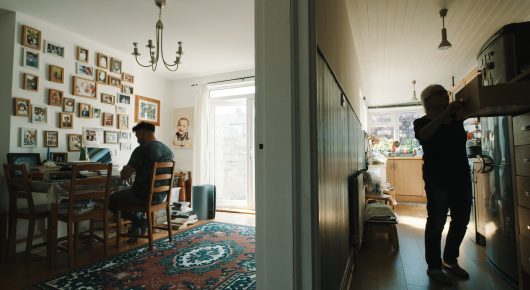 Still from Grappling Grace: a person sits at a table in a room hung with framed photographs, while another person stands at a kitchen counter.