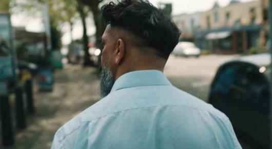 Still from Grappling Grace: a man with dark hair, seen from behind in a light blue shirt, stands on a street.