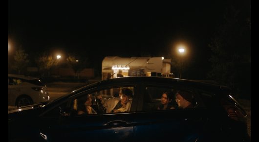 Still from Green Grass: a car is parked at night with people inside, and a caravan with lit windows and street lights behind it.