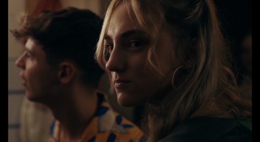 Still from Green Grass: a close-up of a young man and a woman with blonde hair, both looking to the side in dim light.