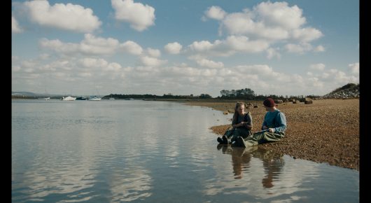 Still from Green Grass: two people sit on the shore of a calm estuary under a blue sky with clouds.