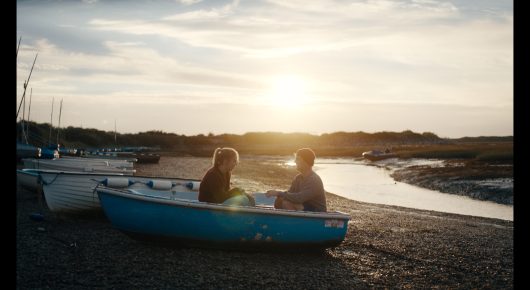 Still from Green Grass: two people sit in a small boat pulled up on a shingle beach at sunset.