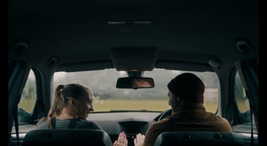 Still from Green Grass: a view from the back seat of a car of two people in the front seats, with a green field through the windscreen.