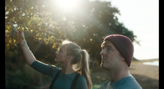 Still from Green Grass: a woman with a ponytail points up at the trees while a man in a burgundy beanie looks the same way, in warm backlight.