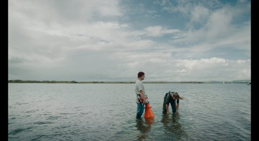 Still from Green Grass: two people wade in shallow water, one bending over, under a cloudy sky.