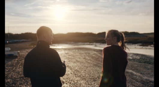 Still from Green Grass: two people stand side by side on the shore at sunset, seen from behind.