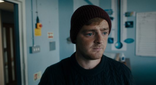Still from Green Grass: a man in a burgundy beanie and dark jumper stands in a blue-tiled kitchen, looking at the camera.