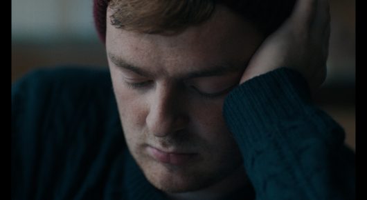 Still from Green Grass: a close view of a man in a dark knitted jumper, his face resting against his hand and his eyes lowered.