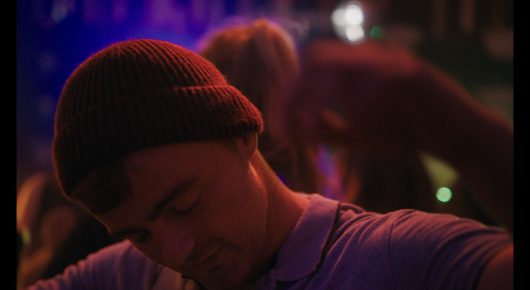 Still from Green Grass: a man in a burgundy beanie looks down in a room lit with purple and red lights, with blurred people behind him.