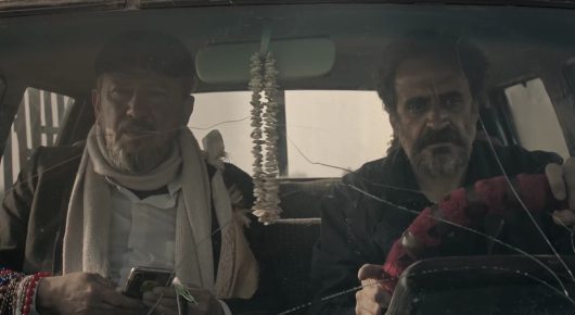 Still from Harsh Land, Distant Sky: two men sit in the front of a vehicle, one in a pale scarf and one gripping the steering wheel.