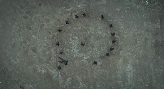 Still from Harsh Land, Distant Sky: an aerial view of a ring of dark figures standing in a circle on pale, textured ground.