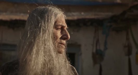 Still from Harsh Land, Distant Sky: a person with long grey hair and a beard in profile, in a dim room with rough walls.