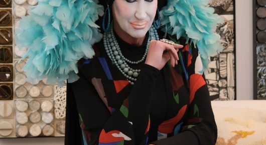 Still from Hunky Jesus: a person in white face make-up and a feathered turquoise collar rests their chin on their hand, wearing a colourful jacket.