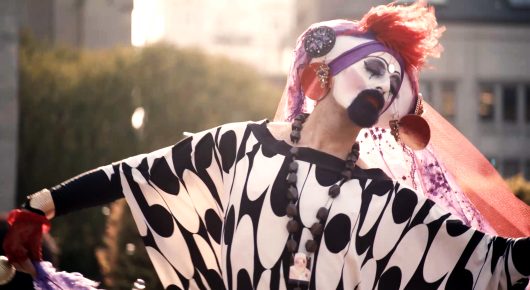 Still from Hunky Jesus: a performer in white face paint, a black-and-white patterned costume and red and purple headwear leans back with arms outstretched in front of a building.