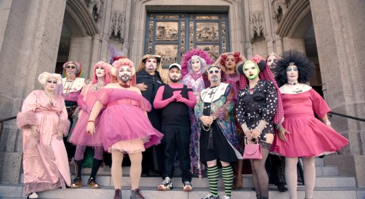 Still from Hunky Jesus: a group of people in bright pink, pale and black costumes and dramatic make-up pose together on the steps of a stone church.