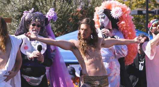Still from Hunky Jesus: a shirtless man with long hair stretches his arms out among people in colourful wigs and costumes, one holding a microphone, at an outdoor event.
