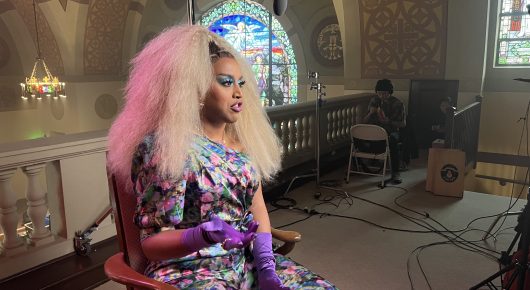 Still from Hunky Jesus: a person with long pale pink hair, a floral dress and purple gloves sits in a chair in a church, with film equipment and cables around.
