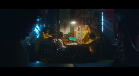 Still from I Love Boosters: several people sit around a table lit by a lamp in a dark, blue-lit room.