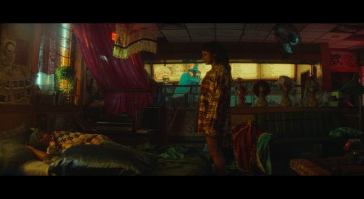 Still from I Love Boosters: a person in a checked shirt stands in a dim, colourful room with pink drapes, while another person lies on a bed in the foreground.