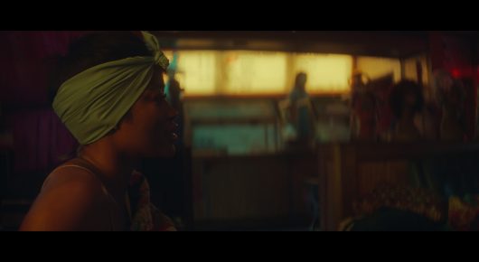 Still from I Love Boosters: a close side view of a woman in a yellow-green head wrap in a warmly lit room.