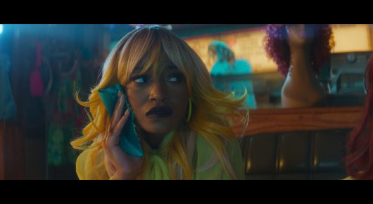 Still from I Love Boosters: a woman with blonde and brown hair holds her hand to her ear in a brightly coloured room with purple and orange lighting.
