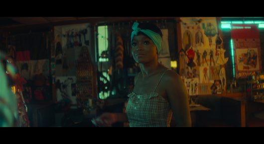 Still from I Love Boosters: a woman in a green head wrap and a sparkly top stands in a room with a wall covered in posters.