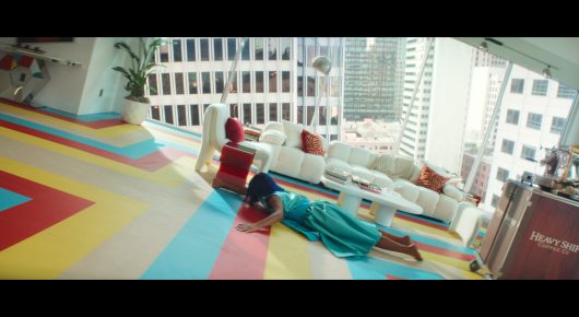 Still from I Love Boosters: a bright living room with a geometric yellow, blue and red floor and a white sofa, where a person lies on the floor, with city buildings outside the window.